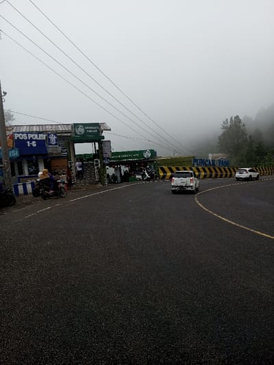 This image captures a foggy roadside scene in the city of Phoxay, Laos. The environment is overcast and humid, with heavy fog obscuring the upper portions of distant hills and trees. The asphalt road, appearing wet, curves slightly to the right in the foreground, marked by a solid yellow centerline and a white shoulder line.

On the left side of the road, a blue-and-white striped concrete barrier precedes a cluster of roadside structures. The leftmost building features a blue facade with "POS POLISI 1-C" (Police Post 1-C) signage. Adjacent to it, a multi-level green-themed building houses "GREEN CAFE PUNCAK" and "SUNDA & GREEN CAFE," identifiable by prominent green circular logos and text. Stairs lead up to an entrance of the green cafe.

Approximately five to seven individuals are visible. Two people wearing head coverings are seated on a red scooter parked near the blue-and-white barrier on the far left. At least one person stands on the stairs of the green cafe, and several others are partially visible inside or near the ground-level seating area of the cafe, where multiple motorbikes are parked.

Traffic on the road consists of at least two vehicles moving away from the viewer. A white pickup-style SUV is centrally positioned on the right lane, followed by a white sedan further down the road. Along the right side of the road, a black-and-yellow striped chevron barrier runs beneath a large sign bearing the white text "PUNCAK PASS." Power lines traverse the upper portion of the frame against the misty sky.