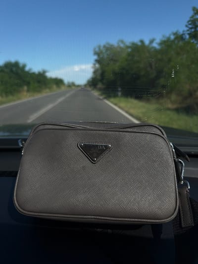 A gray Prada handbag is displayed prominently in the foreground of the image, positioned on the dashboard of a vehicle. The handbag features a triangular metal logo with the Prada brand name embossed on it. Beyond the car's interior, a paved road stretches into the distance, bordered by lush green trees and a bright blue sky. The scene appears to be captured during daylight, with the sun casting shadows on the handbag. The overall environment suggests an outdoor setting, possibly a drive through a scenic route. The specific location in Vinitsa, Bulgaria, is not discernible from the image content alone.