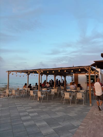 This image depicts an outdoor coastal dining establishment, likely a beach bar or restaurant, at dusk in Sagunto, Spain. The scene features a paved patio area with numerous tables and chairs situated directly adjacent to a beach and the ocean.
A large wooden pergola structure covers a significant portion of the seating area, strung with multiple sets of warm-toned string lights in wicker or woven shades, which are illuminated. To the right, under a similar but separate wooden structure, is a service counter or bar area with a white door and wooden paneling.
Approximately 15-20 individuals are visible throughout the establishment. Many are seated at tables under the pergola, engaged in social interactions, dining, or drinking. One table in the foreground, closer to the ocean, has a pitcher with an orange beverage and glasses. In the far foreground, a man with a shaved head, wearing a white t-shirt with patterned sleeves and dark shorts, walks away from the viewer towards the right side of the frame. Other patrons are standing or moving near the bar area.
The ground is paved with large, dark grey rectangular pavers, arranged in a grid pattern. A strip of reddish-brown pavers forms a border along the right edge of the paved area. Beyond the seating area, a rocky embankment separates the patio from the sandy beach. The ocean stretches into the distance, exhibiting calm, light blue waters with minimal waves. The sky above is a clear, pale blue with subtle cloud formations, gradually transitioning to a faint pinkish hue near the horizon, indicative of twilight conditions. The overall illumination is a combination of natural fading daylight and the warm glow from the artificial string lights.