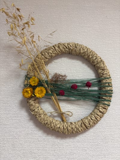 A circular wreath crafted from braided brown paper, approximately 12 inches in diameter, is displayed against a textured white wall. The wreath is adorned with a collection of dried botanical elements. To the left, a cluster of dried grass stalks with seed heads rises from the wreath. Three small, bright yellow dried flowers are also attached to this side. Across the center of the wreath, several strands of dark green yarn are wrapped horizontally. Interspersed with the yarn are three small, dark red dried globe amaranth flowers and a sprig of dried brown foliage. A thin piece of twine is tied in a bow on the lower left portion of the wreath, securing some of the dried stems. The overall impression is a rustic, handmade decorative item. The location is described as Ветрынскі сельскі Савет, Belarus.