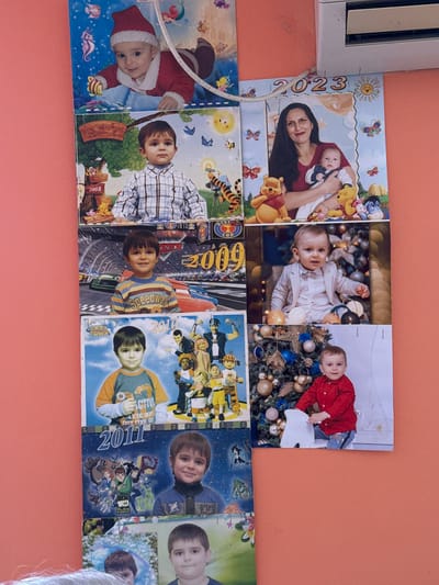 This media file displays a collection of photographs arranged on an orange wall. The photos are printed on what appears to be a calendar or photo album pages, with dates such as 2008, 2009, 2010, 2011, and 2023 visible.

The subjects in the photos are primarily children and babies, with one photo showing a woman holding a baby. Many of the children are depicted in festive or themed settings, including Christmas decorations and what appears to be a race track. Characters from the children's show "Ben 10" are featured in one of the photos. The children's expressions range from curious to joyful.

Objects visible include Winnie the Pooh characters, balloons, Christmas ornaments, and a race car. The setting appears to be indoors, likely a home in Vinarsko, Bulgaria, given the provided location context. The overall mood of the photos is cheerful and celebratory, capturing milestones and memories of childhood. The orientation of the photos is mixed, suggesting they may have been hung at different times or as part of a decorative display.