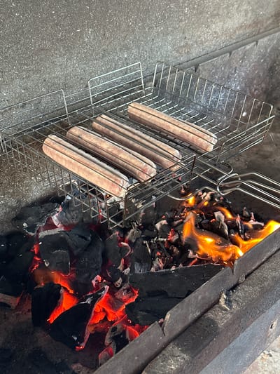 A photograph taken at an elevated angle depicts four sausages cooking in a wire grilling basket positioned above hot coals and flames. The sausages are pale in color and evenly spaced within the basket. The grilling basket is made of thin metal wires. Flames are visible rising from the hot coals, with the fire contained within a rectangular fire pit or grill. The setting appears to be outdoors, with a concrete surface visible to the left of the grill. The image does not contain any people or elements indicative of the city of Kuleshovka, Russia.