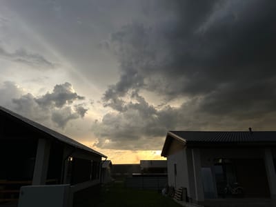 The image depicts a residential area under a dramatic, overcast sky with patches of sunlight breaking through. The scene features several houses with dark roofs, some in various stages of construction or renovation. In the foreground, two houses are visible, one on the left with an extended covered porch, and another on the right with a garage-like entrance. A bicycle is parked near the house on the right. The ground is covered in grass, and there are some construction materials and tools scattered around. The sky is filled with heavy, dark clouds, suggesting an approaching storm or recent rainfall, with shafts of light illuminating the clouds in the distance. The overall atmosphere is somber and moody.