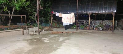 The image depicts an outdoor concrete area at night in Gurakh, Tajikistan, possibly a backyard or courtyard. A corrugated metal roofed structure with decorative iron posts stands in the background, illuminating a section of the scene. Laundry, consisting of several items including a towel and a patterned cloth, is hung on a clothesline suspended between two poles. A broom lies on the ground in the foreground, aligned horizontally. To the left, a simple metal frame structure with a grey plastic crate is visible. The surrounding area is overgrown with trees and vegetation. The ground appears damp, with puddles and scattered leaves.