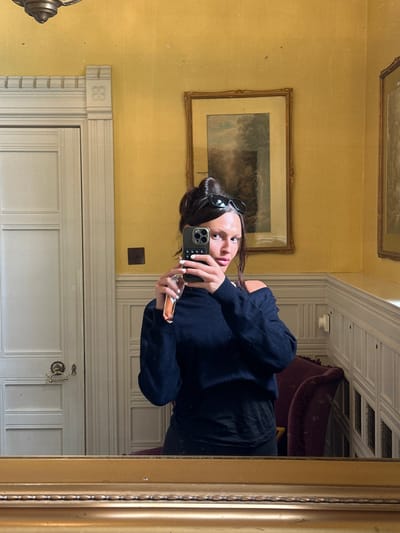 A young woman with dark hair styled in a bun and sunglasses perched on her head takes a selfie in a mirror. She is wearing a dark blue off-the-shoulder sweater and black pants. Her left hand holds a smartphone, and her right hand is extended, holding a small, rectangular object, possibly a candy bar. The background shows a room with yellow wallpaper, a framed painting on the wall, and a white door with ornate molding. The mirror has a thick, golden frame. The scene appears to be an interior of a house.