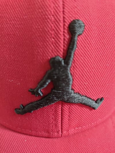 The image is a close-up shot of a red baseball cap with a black Jumpman logo embroidered on it. The logo depicts a silhouette of a person in mid-air, likely a basketball player, with their arms and legs extended. The cap is made of a textured fabric, with visible stitching lines forming a pattern on its surface. The lighting suggests an indoor setting, possibly a room with soft lighting. There are no visible text elements in the image. The focus is solely on the cap and its logo, with the background blurred. The scene is static, with no activity taking place. The image provides a detailed view of the embroidery and the material of the cap.