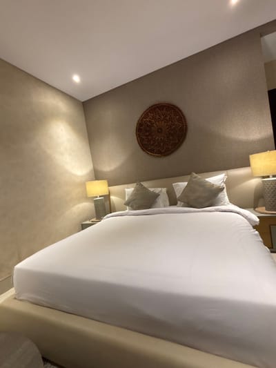 A static interior photograph displays a bedroom located in Batuan, Indonesia. The central subject is a large bed featuring a light-colored upholstered frame, meticulously made with white linens, including a fitted sheet and duvet. Four pillows are arranged against a light-colored, padded headboard: two white standard pillows supporting two smaller, square, light-brown decorative pillows. On either side of the bed, identical light-colored bedside tables are positioned, each holding a matching lamp. These lamps have textured grey bases and cylindrical, warm-toned fabric shades, emitting a soft, ambient light. The wall immediately behind the bed exhibits a light-brown textured finish, and centrally mounted above the headboard is a circular, intricately carved dark wooden medallion. The adjacent left wall presents a lighter beige, textured finish. The white ceiling is equipped with recessed spotlights providing overhead illumination. No individuals or dynamic actions are present within the scene.