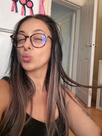 In a room that appears to be a home in Bridlington, United Kingdom, a woman is taking a selfie. She is wearing eyeglasses with tortoise-shell frames and a septum piercing. Her lips are pursed as if for a kiss, and she has long, dark hair with streaks of lighter brown and pink. The background shows a white door with silver handles and a lighter-colored wall. There are also three award ribbons hanging on the wall above her head. The floor is a wooden laminate.