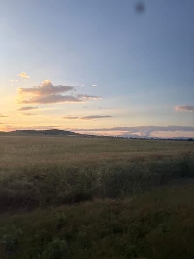 The image displays a landscape scene at sunset or sunrise, located in the city of Svoboda, Bulgaria. The sky occupies the upper two-thirds of the frame, transitioning from a warm golden-orange hue on the left, where several bright, illuminated cumulus clouds are present, to a cooler blue and light gray on the right. A prominent, dark, circular lens flare or smudge is visible in the upper-center sky. The horizon features a dark, undulating hill or low mountain range covered in dense vegetation, silhouetted against the sky. Further to the right, more distant, hazy mountains are faintly visible. The lower third of the image is dominated by a vast field of tall, muted-colored grasses or crops, appearing in shades of dark green, brown, and ochre due to the low light, with the very bottom exhibiting denser, darker foliage. No people, animals, or artificial structures are discernible within the scene.