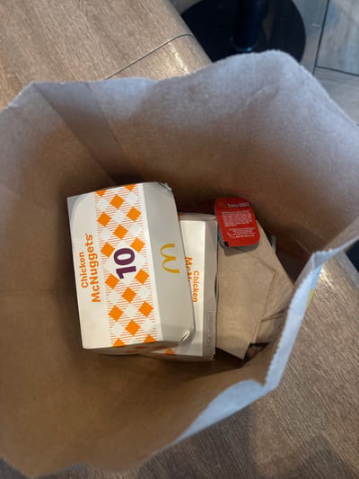 The image shows a McDonald's paper bag with two boxes of Chicken McNuggets, one with the number 10 printed on it, a packet of Salsa BBQ sauce, and paper napkins inside. The bag is sitting on a wooden surface. A blurry black object is partially visible in the background to the left. There is no indication this image was taken in San Buenaventura, Mexico.