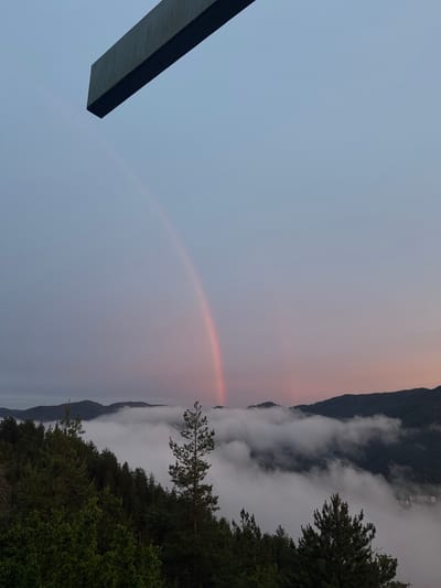 The image shows a natural landscape, likely taken from an elevated viewpoint, in Strazha, Bulgaria. The scene is dominated by a vast expanse of sky and a band of soft, pinkish clouds tinged with the last light of day, suggesting either dawn or dusk. A faint rainbow arcs across the sky, indicating recent rainfall or atmospheric moisture.
Below the sky, a dense layer of white clouds partially obscures a mountainous terrain. The mountains appear as dark, silhouetted forms rising from the cloud cover. The upper slopes of these mountains are covered in dark green forests. In the foreground, on the right side of the image, the dark green foliage of trees, likely conifers and deciduous shrubs, is visible, suggesting a wooded area at the viewpoint.
On the left side of the image, a large, dark, rectangular object juts into the frame from the bottom left corner. Its surface appears to be dark and possibly metallic or stone, with a subtle texture indicating it might be part of a structure, a sculpture, or a viewing platform.
The overall mood is serene and slightly mystical, with the soft colors of the sky, the ethereal clouds, and the subtle rainbow creating a sense of peace and wonder. The time of day is likely early morning or late evening due to the soft lighting and the presence of the rainbow and the atmospheric haze. There is no visible text in the image.