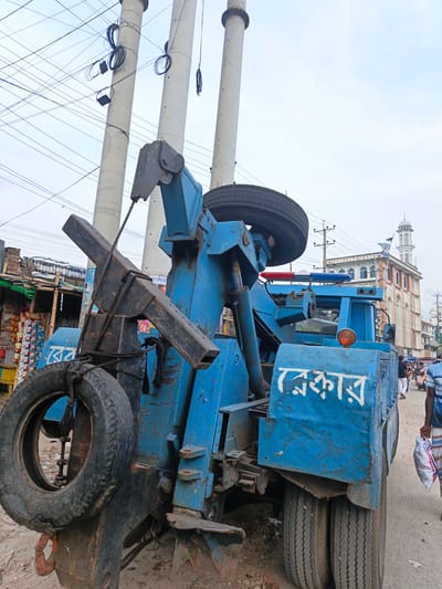 The image depicts a street scene in Savar, Bangladesh, dominated by a large blue tow truck. The truck is parked on a dirt and asphalt road, with several concrete utility poles and numerous overhead wires creating a dense network above. On the left side of the image, a small shop with goods displayed can be seen. In the background, a mosque with a white minaret is visible. A few people are present on the street, with one individual in the foreground on the right carrying a white plastic bag. The tow truck features a prominent boom mechanism with a spare tire attached. White Bengali script is visible on the side of the truck, appearing to say "রেকার" which translates to "wreckers." The sky is overcast.