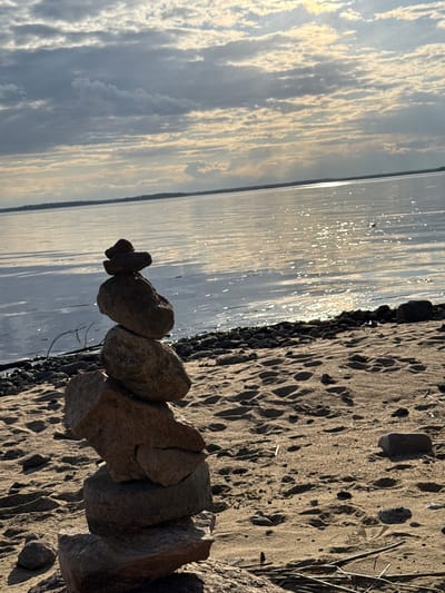 The image shows a stone cairn built on a sandy beach next to a body of water, likely a lake or river. The water reflects the sky, which is filled with clouds. The location is likely not Yasenevo, Russia, as it is a residential area within Moscow and is not known for bodies of water or beaches.