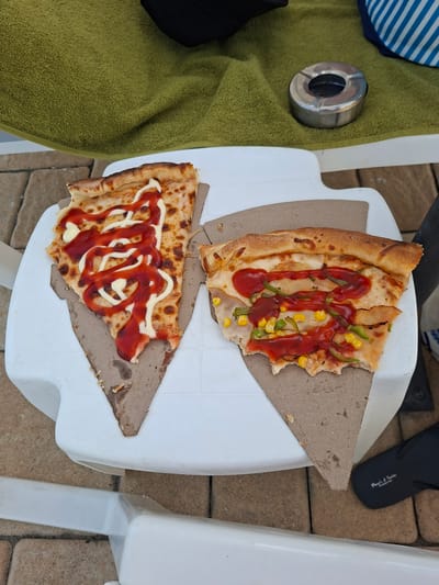 Two slices of pizza, each with visible toppings of ketchup, mayonnaise, corn, and green bell peppers, are placed on a white plastic surface. One slice is topped with a swirled pattern of ketchup and mayonnaise. The other slice appears to have a meat topping underneath the ketchup, corn, and green peppers. A green towel is visible in the background, along with a silver ashtray and a pair of black flip-flops. The scene is set outdoors on a brick patio, suggesting a casual dining or relaxation setting. The presence of the towel and ashtray implies an outdoor recreational area, possibly near a pool or beach.