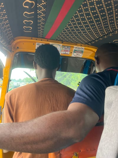 The image, taken in Karmajiji, Nigeria, shows the interior of a yellow auto-rickshaw, likely during daylight hours given the bright lighting. Two individuals are visible. The person in the foreground, wearing an orange t-shirt, is seated and facing away from the camera, looking out the window. Their right arm is resting on the orange seat. Behind them, another person, viewed from the back, is wearing a dark blue t-shirt with red and white trim, and a white fabric is draped over their shoulder.
The interior of the auto-rickshaw features a black fabric panel with a repeating diamond pattern and a Gucci logo emblazoned in gold. There are also vertical green and red stripes on the fabric. Several stickers are affixed to the yellow panel of the auto-rickshaw, including one that reads "KEKE MARTEP 2023 PERMIT" and another with "AREA COUNCIL". The scene appears to be a candid moment, with no clear indication of a specific event. The focus is on the subjects and the details of the vehicle's interior.