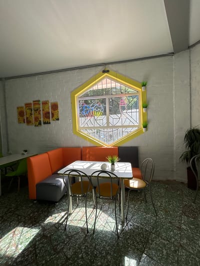 This is an indoor scene of a cafe. A hexagonal window with yellow trim is a central feature, revealing a view of trees and sunlight outside. The window has decorative stained glass depicting ice cream cones. Inside, an orange upholstered booth seating area is arranged around a white-topped table with chrome legs. Two chairs with yellow seats and chrome frames are pulled up to the table. A plant in a white pot sits on the windowsill, and another small plant is on the table. The walls are white brick, and the floor is a speckled green and white terrazzo. The scene is illuminated by bright sunlight casting distinct shadows on the floor. There are no people visible in the image. Based on the limited visual information, it is not possible to definitively identify the specific city or country, and therefore the location in Rodniki, Russia cannot be confirmed.