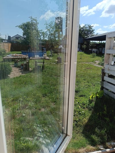 The image captures a sunny day in a rural outdoor setting, possibly a backyard or garden, in Цялушскі сельскі Савет, Belarus. A person is lying on a wooden platform, appearing to sunbathe. In the background, a blue inflatable pool is visible, along with a wooden table and chairs, suggesting recreational activities. A covered structure, likely a shed or carport, stands further back, with wooden fencing and a white pallet structure present. The environment is characterized by green grass, trees, and a bright blue sky with scattered clouds. The scene is viewed through a window, with the frame of the window partially obscuring the view.
