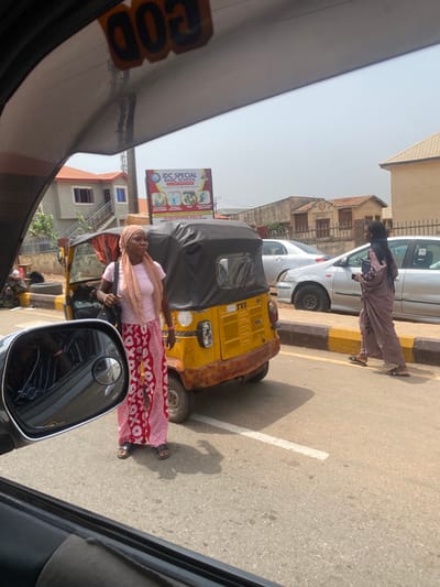 The image shows a street scene in AlakIa, Nigeria. A woman wearing a headscarf, light pink shirt, and red and white patterned skirt stands next to a yellow auto-rickshaw (TVS brand). Another woman in a hijab is walking along the road next to a white car. A sign for "IDC Special Basic School" is visible in the background. The view is taken from inside a vehicle, with parts of the dashboard and side mirror visible.