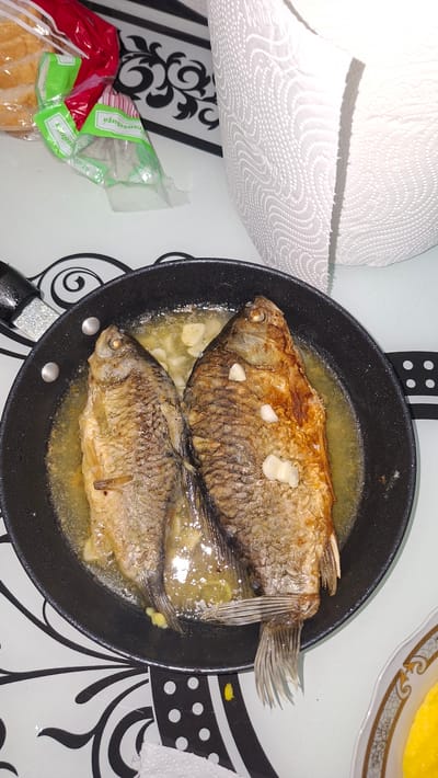The image shows a close-up overhead view of two whole fried fish in a black frying pan. The fish are golden brown and appear to be cooked in oil with some visible garlic pieces. The frying pan is placed on a white surface with a black and white patterned tablecloth. To the left of the pan is a red and green plastic bag, and to the right is a roll of white paper towels. In the upper left corner, a portion of sliced bread is visible within a clear plastic bag. A small decorative plate with a yellow substance is partially visible in the lower right corner. The scene appears to be indoors, likely in a kitchen or dining area. There are no people visible in the image. The location of Voinova, Moldova, is not discernible from the visual elements present in the image.