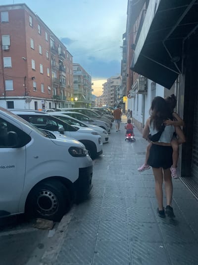 The image depicts a street scene in Sagunto, Spain, at dusk. A woman is seen from behind, carrying a young child on her back. The child is embracing the woman. To their left, a child rides a small, pink electric toy motorcycle. Further down the street, a man in an orange t-shirt and shorts walks away from the camera. Several cars are parked along the side of the street, and buildings with brick facades line the left side of the road. The sky above is overcast with a gradient of colors indicating the setting sun. The sidewalk is paved with rectangular tiles, and a white van is parked in the foreground on the left.