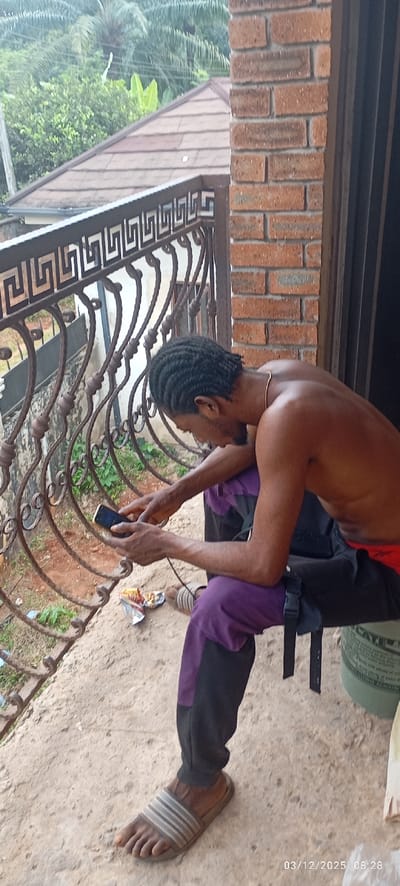 An individual is depicted seated outdoors in Abagana, Nigeria, on a concrete surface, leaning forward and operating a black mobile phone with both hands. The subject is an adult male with short, braided dark hair, wearing a thin gold-colored necklace. He is shirtless and wears purple and black athletic-style pants. A grey and white striped slide sandal is visible on his right foot. An open, crumpled wrapper containing a snack item rests on the concrete near his right foot. To his right, a partially visible green plastic bucket or container supports him. A dark brown metal railing with a Greek key pattern is in the foreground, separating the subject from a vegetated area below. Behind the subject to the right is a reddish-brown brick column or wall. In the background, a dark brown tiled roof is visible beneath a dense canopy of green foliage, including palm trees and banana leaves. The image bears a timestamp of 03/12/2025 08:28.