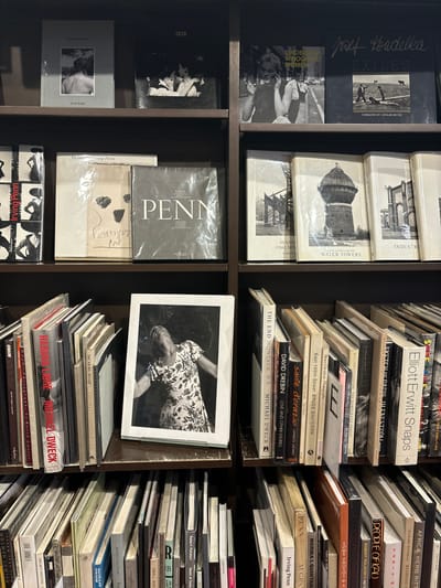 A bookshelf in a bookstore in Chiyoda City, Japan. It contains a selection of hardcover books, most of which are photography books. There are three shelves on the left, with each one holding a few books. On the top shelf, the books have titles such as "Exiles," "Indiser," and "Water Towers." On the second shelf, there are books titled "Endangered Women," "Penn Coleman" and "Izis." The bottom shelf holds one book with the title "Irving Penn." The shelves on the right are packed with books. There is a picture of a woman in a floral dress on the middle shelf, with the words "LOVE AND OTHER STORIES" on the bottom of the frame. The top shelf has books titled "Elliott Erwitt Snaps," "Sketch of Paris in the Storm," and "The Democratic Forest." The shelves below it have books with titles such as "The End Montauk, NY," "Love and Other Stories," "The Sweet Life of the City," "Habana Libre," and "One Dinner."