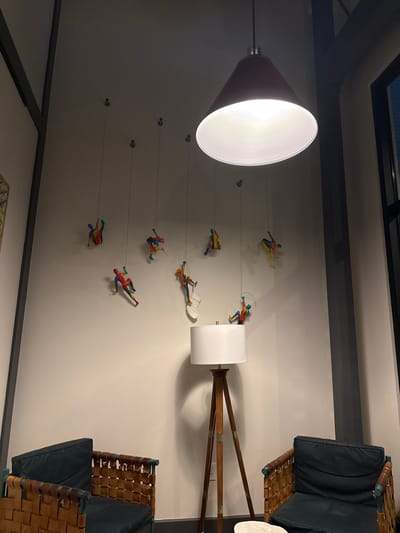 A dimly lit interior showcases a cone-shaped pendant lamp emitting a bright light. On the adjacent wall, a whimsical display features figures suspended by thin wires, seemingly climbing towards a tripod lamp with a cylindrical shade. The scene also includes two armchairs with dark cushions and woven backs, positioned against the wall.