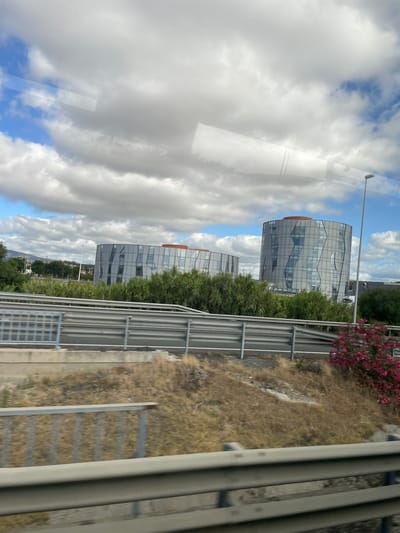 The image, taken from a moving vehicle, shows two modern, circular buildings with glass facades in Los Barrios, Spain. The buildings feature irregular, vertical patterns on their glass exteriors and have orange circular roofs. They are surrounded by lush green foliage. In the foreground, a metal guardrail and fence are visible, along with dry, grassy terrain. The sky is partly cloudy with a mix of white and gray clouds against a blue sky. A lamppost stands to the right of the buildings. The overall scene depicts an urban or industrial area with a focus on contemporary architecture.