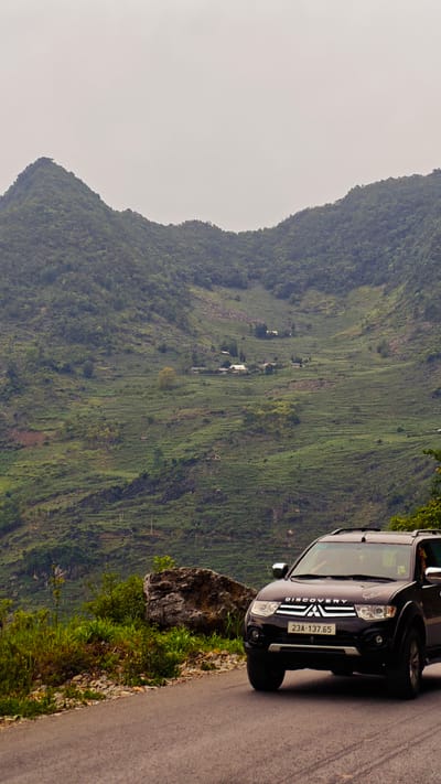An eye-level, medium shot depicts a dark-colored Mitsubishi SUV parked on a paved road in Cán Tỷ Commune, Vietnam, set against a backdrop of verdant, terraced hills and a cloudy sky. The SUV is positioned to the right, with its license plate, “23A-137.65,” visible. A large rock and some shrubbery appear on the roadside beside the vehicle. The terraced hills are densely covered with vegetation, and a small cluster of buildings is visible in the distance. The sky is overcast, casting a muted light over the scene.