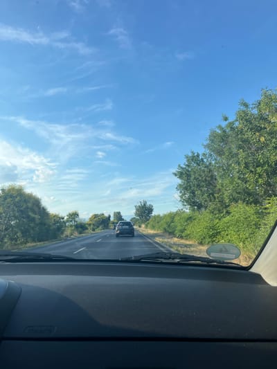 The image shows a view from inside a car driving on a two-lane road in Graf Ignatievo, Bulgaria. The sky is blue with scattered white clouds. On the right side of the road, there is a dense line of green trees and bushes. On the left side, there are also trees and vegetation. The road ahead has two cars visible, one in the distance and one closer, both in the same lane. The dashboard of the car is in the foreground.