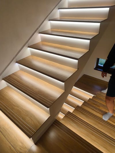 The image depicts a well-lit staircase in an indoor setting. The stairs are constructed with wooden treads and white risers, with integrated LED lighting underneath each tread. The wooden treads feature a grooved design. A person wearing shorts and socks is partially visible on the lower steps, descending the staircase. A small window is present on the right side of the image near the stairs. The location is Mosomishte, Bulgaria.