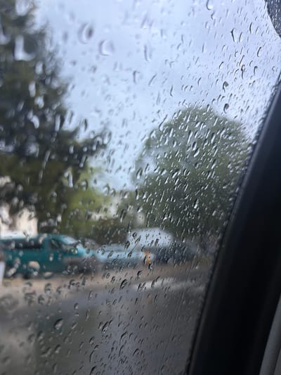 The image depicts a rain-covered window, likely a car window, with an out-of-focus background. The background appears to show trees and buildings. The location in Davidkovo, Bulgaria cannot be confirmed.