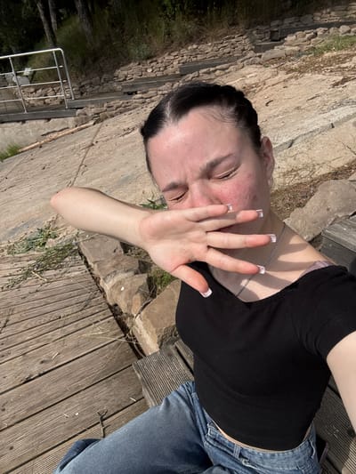 A young woman with dark hair braided and pulled back sits on a wooden bench outdoors, covering her face with her left hand. Her eyes are closed, and her facial skin appears flushed. She is wearing a black cropped t-shirt and blue jeans. Behind her, a stone embankment with steps and a metal railing leads upwards towards trees. The ground is a mix of concrete and wooden planks. There is no indication in the image that this scene is located in Pozdyshki, Russia.
