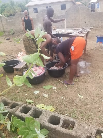 Four individuals are preparing a meal in an outdoor space. Two men and two women are involved in the activity. One man, dressed in a black shirt and camouflage pants, is hunched over, processing meat. A second man, shirtless, is standing behind a table holding a piece of raw meat. One woman is kneeling, processing meat in a black container. A second woman, dressed in orange shorts and a black shirt, is standing nearby, and is processing meat in a separate black container. Several large pieces of meat are visible in black containers along with other butchered meat. The area is primarily dirt with some scattered foliage. There is a low wall made of cinder blocks in the foreground. The location is Pati Ridge, Nigeria, at coordinates (latitude: 7.81, longitude: 6.704).
