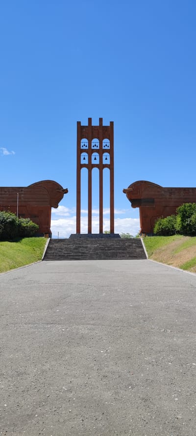 The image captures a monument located in the city of Araks, Armenia. In the foreground, a paved pathway leads towards a wide set of concrete stairs. The stairs ascend to a raised platform where a striking bell tower stands prominently. The tower is made of a reddish-brown material and features multiple arched openings, each containing bells. On either side of the bell tower, symmetrical, large, sculpted figures resembling horse heads, also made of the reddish-brown material, flank the scene. Behind the monument, a clear blue sky with scattered white clouds provides a backdrop. Green grass covers the ground around the base of the monument and the sculpted figures.