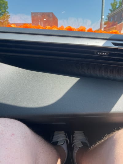 The image shows a person's legs and feet inside a car, viewed from a slightly elevated perspective. The person is wearing white and black sneakers. The interior of the car is visible, with textured plastic surfaces and what appears to be a sun vent. A decorative garland of orange flowers is draped along the edge of the car's window, reflecting the bright blue sky and some buildings and trees outside. Sunlight is casting shadows on the car's dashboard. The scene suggests a sunny day, possibly in the Netherlands given the orange floral decoration, which is commonly associated with national celebrations. The overall impression is one of a casual journey on a pleasant day.