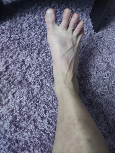 The image shows a close-up, aerial view of a person's bare foot and lower leg resting on a fluffy, purple carpet. The foot is positioned at an angle, with the toes pointing towards the top left of the frame. The skin on the foot and leg appears pale and shows visible veins. The toes are spread slightly, and the toenails are visible. The carpet is textured and appears to be shaggy. A dark object, possibly a piece of furniture, is partially visible in the upper right corner. The lighting suggests an indoor setting. There is no identifiable context or information within the image to associate it with the city of Gorno-Altaysk, Russia.