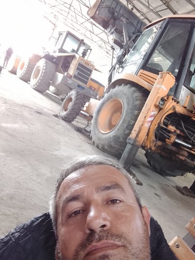 A middle-aged man with graying hair and a stubble beard is taking a selfie. He is wearing a black jacket. Behind him are several pieces of heavy construction equipment, including a backhoe and a front-end loader. The equipment is located indoors, likely in a garage or warehouse with a metal roof structure visible overhead. The location is likely in the city of Sabir Rakhimov, Uzbekistan.
