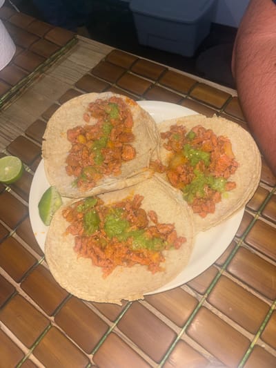 The image shows a plate of three tacos with orange-colored meat, green sauce, and two lime wedges on a wooden table. The location is Buenavista, Mexico. A person's arm is visible in the upper-left corner of the image.