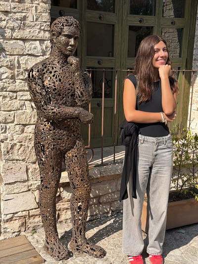 The image shows a woman lying next to a metal sculpture of a person, likely in Domingo Arenas, Mexico. The woman is smiling at the camera, wearing a black top, grey jeans, and red shoes. She has long brown hair and is wearing bracelets on her right wrist. The sculpture is lying on a stone wall, next to the woman. The background includes a green wooden door with dark metal accents, some plants, and a stone patio. The lighting suggests a sunny day.