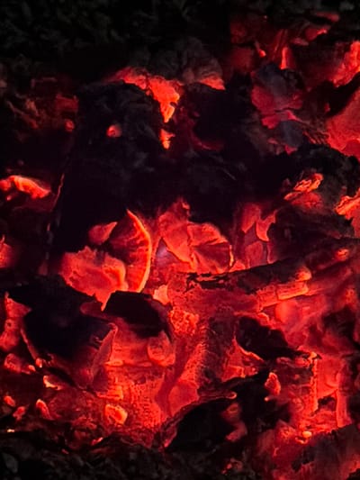 The image is a close-up, high-angle shot of glowing embers and charred wood from a fire. The dominant colors are vibrant reds, oranges, and deep blacks, indicating intense heat. The texture is rough and irregular, with sharp edges and uneven surfaces on the wood fragments. There are no people, animals, or any identifiable objects other than the remnants of a fire. The scene is dark, with the only light source being the embers themselves. The image does not provide any information about a specific location in Zimyonki, Russia, or any context related to its surroundings.