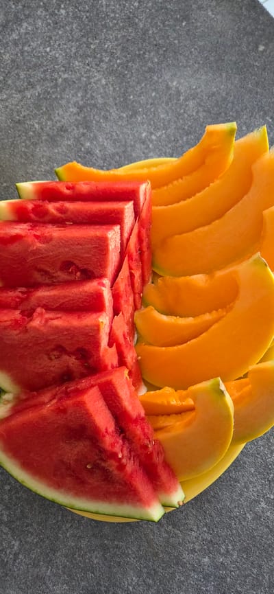 A close-up, vertical, eye-level shot shows sliced watermelon and cantaloupe arranged on a yellow plate. The watermelon slices, a vibrant red with white rinds, are stacked in a stepped pattern on the left side of the plate. The cantaloupe slices, a bright orange with a green-tinged rind, are fanned out on the right side. The plate rests on a dark gray, textured surface, possibly a countertop or table. The lighting suggests an indoor setting or an overcast outdoor environment, casting soft shadows. The image does not contain any specific clues to pinpoint its location within Lignano Sabbiadoro, Lignano, Italy.