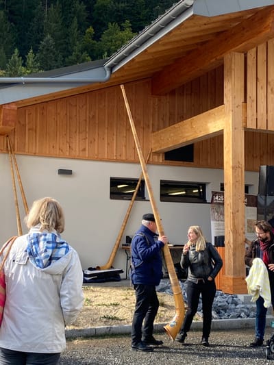 The image captures an outdoor cultural event in Abondance, France, situated in front of a modern building with a forested mountain slope in the background. The building features a light gray lower facade, natural wood plank cladding on the upper story, and exposed timber structural elements, including a prominent vertical beam. A gray gutter system runs along the roofline.
In the mid-ground, a middle-aged male figure, dressed in a dark blue jacket, dark trousers, and a dark cap, stands vertically holding a tall, light-brown wooden alphorn. The alphorn's flared bell end, resting on the ground, exhibits a painted floral motif in blue, pink, and green. He is in conversation with a blonde woman to his right, who wears a black leather jacket, black trousers, and black boots, smiling and gesturing with her right hand. To the left of the man, another identical alphorn stands upright, leaning against the building. Two additional alphorns are partially visible further left, also leaning against the building's facade. A black equipment case or stand is on the ground near the central alphorn, and dark sound equipment is partially visible to the right.
On the far right, a bearded man, partially obscured, holds a yellow and white checkered cloth. In the foreground, a blonde woman, seen from behind, wears a light-colored jacket with a blue and white checkered hood lining and carries a pink-patterned bag over her left shoulder, observing the scene.
The ground consists of light-colored gravel and sparse vegetation. The scene is illuminated by natural daylight.