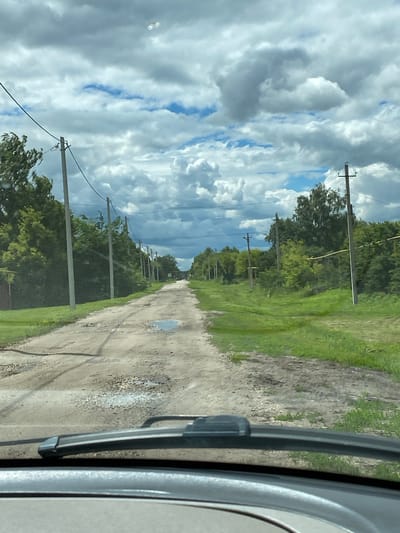 An eye-level, wide-angle shot looks down a rural, unpaved road in Nadezhdino, Russia. The road is rutted and has several puddles, suggesting recent rain. Telephone poles with wires line both sides of the road, interspersed with trees and green grass. The sky is cloudy with patches of blue sky visible. The foreground shows the windshield and wiper of a vehicle.