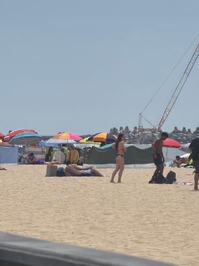 The image depicts a crowded beach scene in Espinho, Portugal, under a clear, pale blue sky. In the foreground, the sandy beach is occupied by several individuals and beach equipment. To the left, a person is lying on their stomach on a towel, wearing a blue and red bathing suit. To the right, another person is lying on their back, facing left, wearing dark swim trunks.
Mid-ground, a group of beach umbrellas are set up. A prominent orange and pink umbrella is positioned behind the person lying on their back. Further left, a red and blue striped umbrella is visible. Several tents or windbreaks are also present, providing shade and privacy for beachgoers.
In the background, a large construction crane with an orange boom is visible against the sky. The crane is situated behind a line of large, grey, concrete wave-breaking structures that extend into the ocean. People are scattered throughout the scene, some relaxing under umbrellas, others walking or standing. A woman in a black bikini is standing in the middle ground, facing right, and a man in dark swim trunks is standing to her right, facing left, with his hands on his hips. Bags and towels are visible on the sand near them. The overall impression is one of a typical busy summer day at the beach.