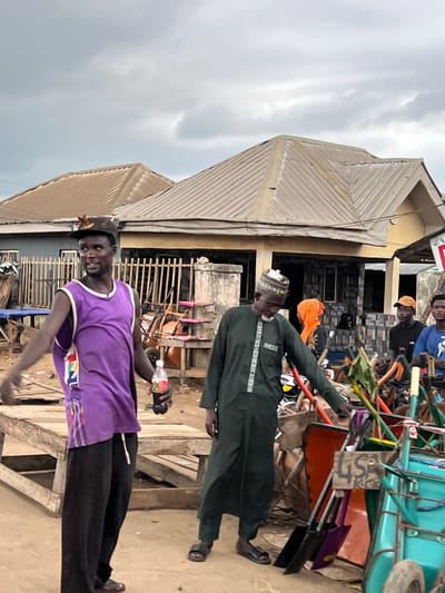 In Oko Amakom, Nigeria, a medium shot captures a street scene with several people and an outdoor market. In the foreground, a man wearing a purple tank top and black pants stands holding a bottle of soda. To his right, another man in a dark green robe and cap examines tools. Behind them, various market stalls and goods, including shovels and wheelbarrows, are visible. Two buildings with corrugated metal roofs form the background. The sky is overcast with a few visible clouds.