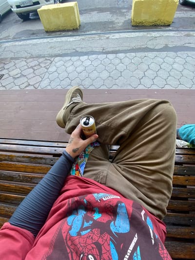 The image depicts a person sitting on a wooden bench in what appears to be an outdoor urban setting. The person is wearing brown corduroy pants, a red t-shirt with a graphic print, and a dark long-sleeved undershirt. They are holding a can of what appears to be a beverage in their right hand. The background shows a paved area with a drainage grate, a white vehicle with a license plate that reads "P051VT 27", and two large yellow concrete blocks. The overall environment suggests a public space, possibly a street or a plaza, in the city of Mayak, Russia.