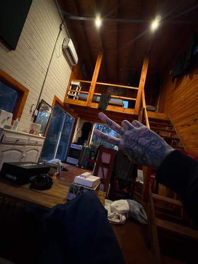In Dique Luján, Argentina, a person's tattooed hand is in the foreground, displaying a peace sign gesture. The hand is positioned in front of a rustic, wooden interior. The background reveals a two-story cabin-like structure with exposed wooden beams and paneling. A loft area is visible with a wooden staircase leading up. The room contains furniture, including a white cabinet with drawers, a mirror, and a kitchen stove. An air conditioning unit is mounted on a light-colored wall. The lighting suggests it is evening or nighttime.