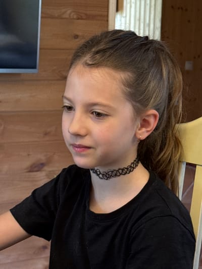 A young girl with brown hair pulled back in a ponytail and a black choker necklace is looking to her left. She is wearing a black t-shirt. The background consists of a wooden wall with horizontal planks and a blurred yellow chair to the right. To the left, a dark television screen is visible. The image appears to be taken indoors, possibly in a rural setting or a cabin, given the wooden interior. There is no specific information to pinpoint the location within Zassovskaya, Russia.