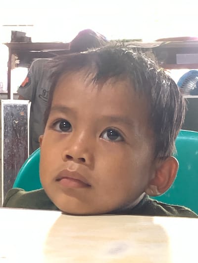 A close-up portrait of a young child with dark hair and eyes, resting their chin on a white surface, possibly a table. The child is wearing a dark green shirt. Behind the child, a blue plastic chair is visible. In the blurred background, parts of a room setting can be discerned, with an unidentified person wearing a grey shirt with what appears to be an emblem. There's also a glimpse of a fan. The exact location in Samarinda, Indonesia, cannot be determined from the image.